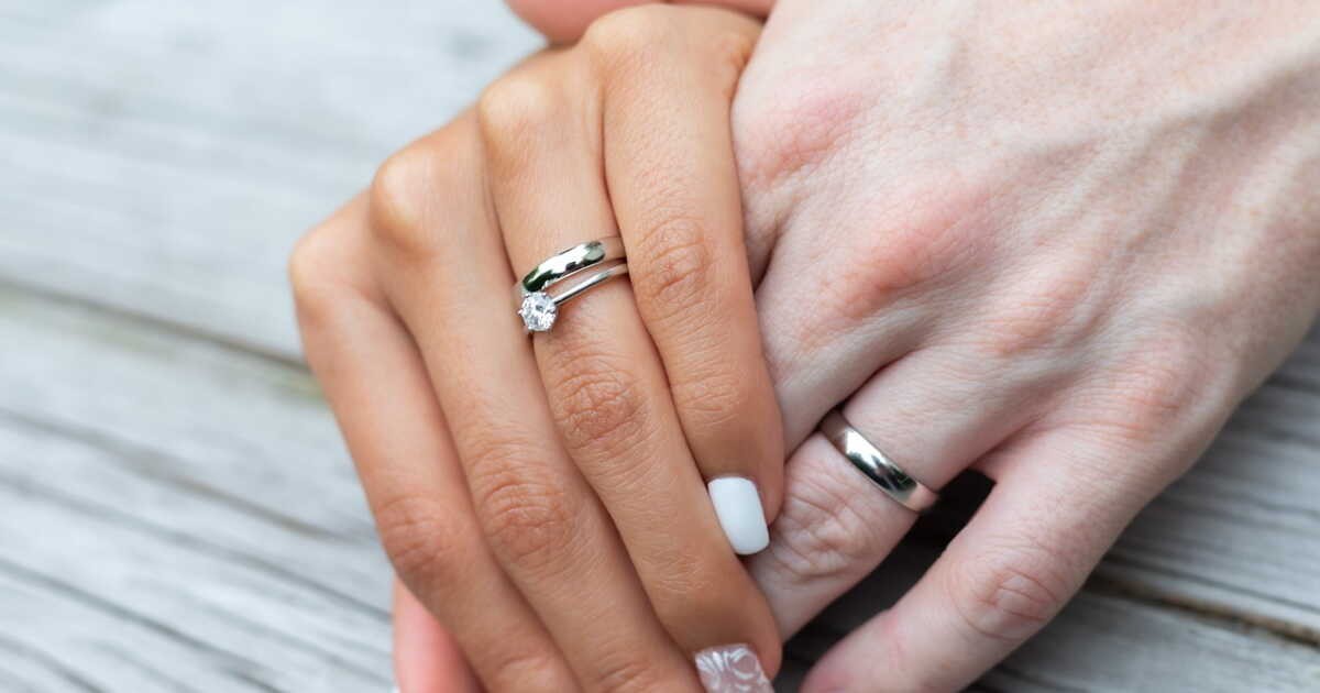 Gros plan sur deux mains enlacées portant des alliances en argent, l’une ornée d’un solitaire diamant à l’annulaire droit, symbole d’amour, d’engagement et de la signification culturelle de l’alliance main droite.